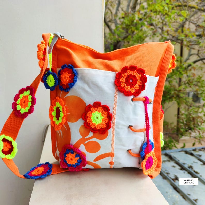 K&ouml;nnte beinhalten: Eine orangefarbene Umh&auml;ngetasche mit einer wei&szlig;en Frontplatte mit orangefarbenen Blumenmustern. Die Tasche ist mit bunten, geh&auml;kelten Blumenapplikationen in Rot, Blau und Gelb verziert. Der Riemen hat auch geh&auml;kelte Blumen.