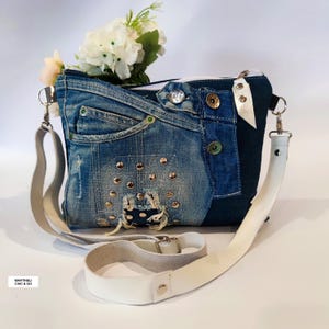 May include: A denim crossbody bag with a white leather strap. The bag is made from recycled blue jeans and features metal studs, buttons, and a zipper closure. A small bouquet of white flowers sits on top.