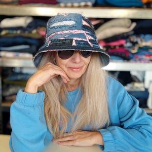 以下が含まれることがあります： パッチワークデザインのデニムバケットハット。青いデニムとフローラル生地のアクセントが特徴です。サングラスと青いスウェットシャツを着た人が着用しています。「MARTINELI CHIC & GO」の文字が見えます。