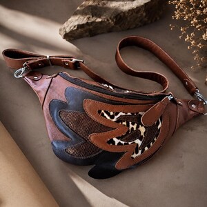 May include: A brown leather fanny pack featuring a zippered main compartment and an adjustable strap. The front is decorated with layered appliques in dark brown, textured bronze, and a leopard print pattern, secured with silver-toned metal hardware.