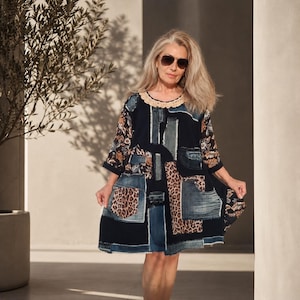 Op de afbeelding: Een knielange jurk met een patchworkontwerp van donkerblauwe denim, stof met luipaardprint en bloemmotieven. Het kledingstuk heeft driekwartmouwen, een ronde halslijn met een beige gehaakte rand en meerdere zichtbare zakken.