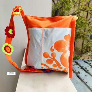 K&ouml;nnte beinhalten: Orangefarbene Umh&auml;ngetasche mit wei&szlig;er Frontplatte mit gro&szlig;em orangefarbenem Blumendesign. Die Tasche hat einen orangefarbenen Riemen, der mit bunten geh&auml;kelten Blumen verziert ist. Der Text "MARTINELI CHIC & GO" ist auf dem Riemen zu sehen.