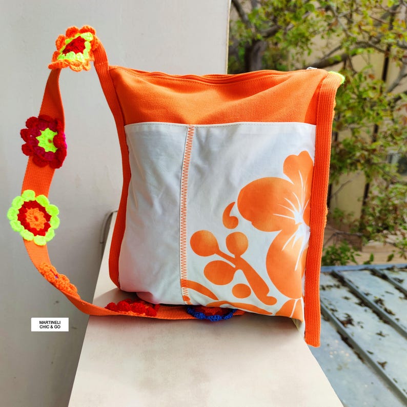 K&ouml;nnte beinhalten: Orangefarbene Umh&auml;ngetasche mit wei&szlig;er Frontplatte mit gro&szlig;em orangefarbenem Blumendesign. Die Tasche hat einen orangefarbenen Riemen, der mit bunten geh&auml;kelten Blumen verziert ist. Der Text "MARTINELI CHIC & GO" ist auf dem Riemen zu sehen.