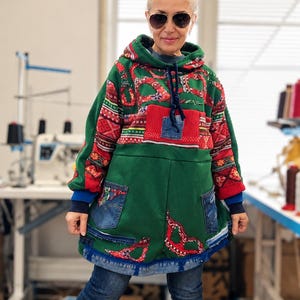 Op de afbeelding: Een groene hoodie met een patchworkontwerp van rood-wit patroonstof, donkerblauwe spijkerzakken en een gerafelde spijkerzoom. Het kledingstuk is voorzien van een capuchon met trekkoord en lange mouwen met geribbelde boorden.