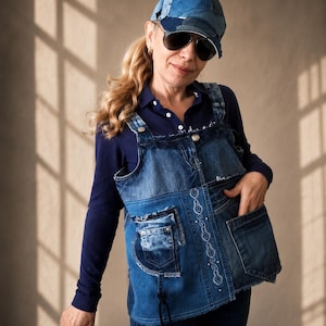 Peut inclure: Un gilet en jean patchwork composé de différentes nuances de bleu, avec des bords effilochés et des broderies blanches. Le gilet présente plusieurs poches et une fermeture à boutons, porté sur une chemise bleu marine à manches longues.