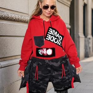 Può includere: Una felpa rossa con cappuccio dotata di taschino sul petto in denim nero, una toppa nera con la scritta bianca "MODE CAME" e una toppa rettangolare con paillettes. La parte inferiore è in denim mimetico nero e grigio con tiretti rossi.