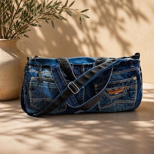 Könnte beinhalten: Eine zylindrische Reisetasche aus dunkelblauem Jeansstoff mit ausgefransten Kanten. Die Tasche verfügt über schwarze Nylongurte, silberfarbene Metallbeschläge und einen kleinen gestickten Aufnäher auf der Vordertasche mit einem geflügelten Emblem.
