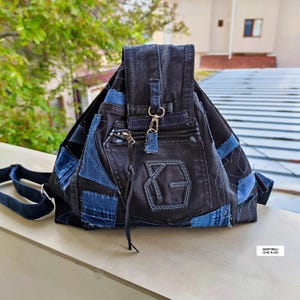Peut inclure: Un sac à dos en jean noir avec des patchs en jean bleu et une fermeture à rabat. Le sac à dos comporte une poche zippée et un fermoir argenté. Le sac a des bretelles réglables et un design cousu. Le texte "MARTINELI CHIC & GO" est visible.