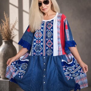Op de afbeelding: Een knielange tuniekjurk met een patchworkontwerp van blauwe denim, rood-wit ruitpatroon en witte stof met blauwe en rode geometrische patronen. Het kledingstuk heeft korte mouwen met blauwe kanten randen, een ronde hals en twee voorzakken.