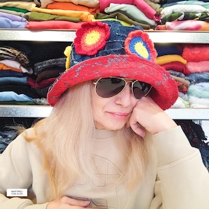 Może przedstawiać: Czapka typu bucket hat wykonana z ciemnoniebieskiego dżinsu, z szerokim rondem z czerwonej koronki oraz żywymi, wielokolorowymi szydełkowanymi aplikacjami kwiatowymi w odcieniach żółci, pomarańczu i błękitu. Noszona z przyciemnianymi okularami.