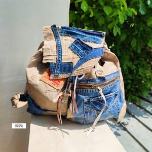 May include: A beige and blue denim backpack with a patchwork design. The backpack features a front pocket with a zipper and a red tag. The bag has a drawstring closure and leather accents. The backpack is made of recycled denim.