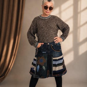 Op de afbeelding: Een vrouw draagt een bruine trui met een wit en zwart patroon, een denim patchwork rok met een bloemenontwerp, zwarte leggings en bruine laarzen. De rok heeft verschillende denim patches en een bloemen applicatie. De vrouw draagt een zonnebril.