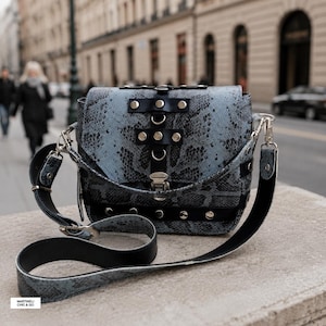 Peut inclure: Un sac à bandoulière structuré avec un motif peau de serpent bleu et noir, agrémenté de lanières en cuir noir, de clous métalliques argentés et d'un fermoir central. Le sac comprend une bandoulière noire amovible et une étiquette MARTINELLI CHIC & GO.