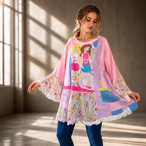 Op de afbeelding: Een roze tuniek met lange mouwen en een patchwork-ontwerp. De tuniek heeft een afbeelding van twee cartoonmeisjes, mouwen met bloemenpatroon en een kanten rand. Het model draagt een blauwe spijkerbroek.
