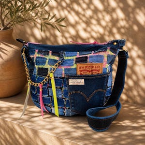 Puede incluir: Un bolso de hombro de mezclilla tipo patchwork con un diseño tejido de cuadrados de tela azul con detalles en rosa neón y amarillo. Incluye un bolsillo frontal con una etiqueta de Levi Strauss & Co., una cadena dorada, borlas de cuero neón y una correa de mezclilla.