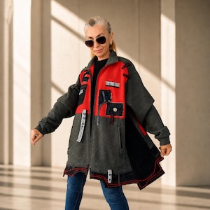 May include: A charcoal gray zip-up jacket featuring vibrant red panels on the chest and back. The jacket includes multiple black patch pockets, decorative black and white striped fabric accents, and a black hemline with red stitching details.