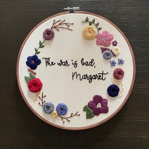 May include: Embroidered hoop art featuring a floral wreath and the text "The war is bad, Margaret".