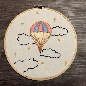 May include: A white embroidery hoop featuring a hand-stitched design of a hot air balloon with pink, blue, and beige stripes, floating in the sky with clouds and stars.