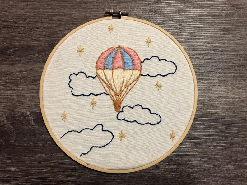 May include: A white embroidery hoop featuring a hand-stitched design of a hot air balloon with pink, blue, and beige stripes, floating in the sky with clouds and stars.