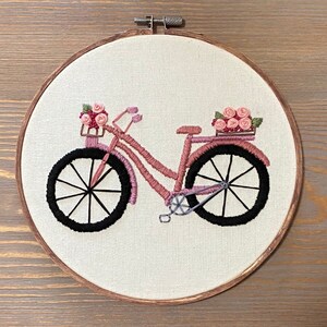 May include: Embroidered pink bicycle with a basket of pink flowers on a white background. The bicycle has black wheels and a brown wooden frame.