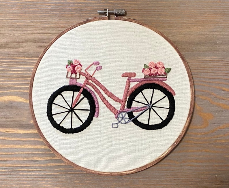 May include: Embroidered pink bicycle with a basket of pink flowers on a white background. The bicycle has black wheels and a brown wooden frame.