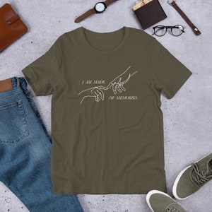 May include: Olive green t-shirt with white line art of two hands nearly touching, and the text "I AM MADE OF MEMORIES". The shirt is short-sleeved and made of a soft material.