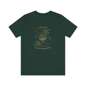 Pode incluir: Uma camiseta verde escura com uma ilustração dourada de duas mãos segurando uma chama. O texto "I was made to pass through fire." está impresso na camiseta.