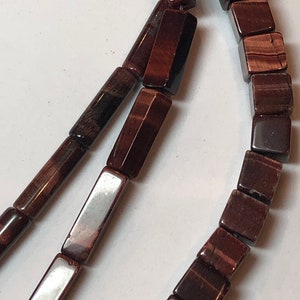 May include: A close-up of a necklace made of dark red-brown beads. The beads are a mix of square and rectangular shapes, strung together. The necklace is displayed against a white background.