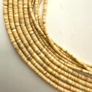 May include: Close-up of a necklace made of small, round, beige shell beads. The beads are strung together in multiple strands, creating a layered effect. The necklace has a natural, earthy aesthetic.