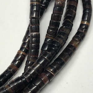 May include: Close-up of a necklace made of dark brown, cylindrical beads. The beads have a polished, glossy finish and are strung together to form multiple strands. The beads are a mix of dark and light brown.