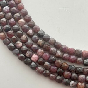 May include: Strands of faceted, dark red-brown gemstone beads. The beads have a square shape with multiple facets, creating a sparkling effect. The beads are strung together, forming multiple rows. The background is a plain white surface.