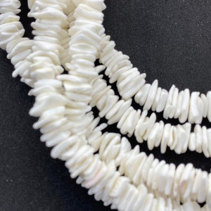 May include: Close-up of a white shell necklace. The necklace is made of small, irregularly shaped shell pieces strung together. The shells have a slightly glossy finish and are arranged in multiple strands. The background is a solid black.