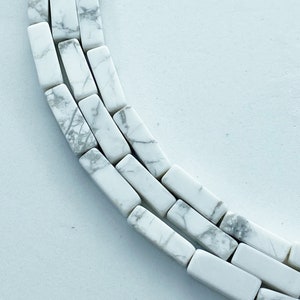 May include: Close-up of a necklace or strand of white rectangular beads with gray marbling. The beads are arranged in a curved pattern against a white background. The beads are approximately 0.75 inches long.