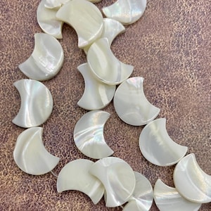 May include: A collection of iridescent, crescent-shaped mother-of-pearl beads. The beads are a creamy white color and have a subtle shimmer. They are scattered on a textured, brown surface.