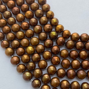 May include: Close-up of strands of lustrous, round, bronze-colored pearls. The pearls have a smooth, reflective surface with hints of gold and green iridescence. The beads are strung together, showcasing their uniform size and shape.