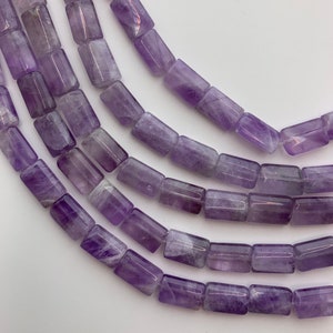 May include: Close-up of strands of amethyst gemstone beads. The beads are rectangular, with a polished finish, and vary in shades of purple. The beads are strung together, creating a flowing, curved line against a white background.