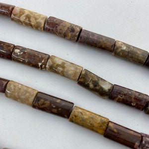 May include: Close-up of strands of natural stone beads. The beads are cylindrical, with a mix of brown, tan, and burgundy colors. The beads have a polished finish and are strung together, ready for jewelry making.