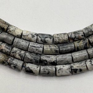 May include: Close-up of a strand of gray and white cylindrical beads. The beads have a marbled appearance with dark gray and black veining. The beads are strung together, forming a curved shape.