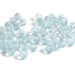 May include: A collection of light blue, faceted beads scattered on a white surface. The beads are round with multiple facets, creating a sparkling effect. These beads are suitable for jewelry making and craft projects.
