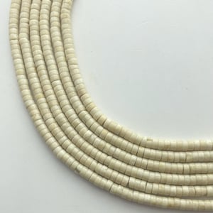 May include: Close-up of several strands of off-white, cylindrical beads. The beads are uniform in size and shape, with a slightly textured surface. The strands are arranged in a curved pattern against a white background, suggesting a necklace or similar jewelry.