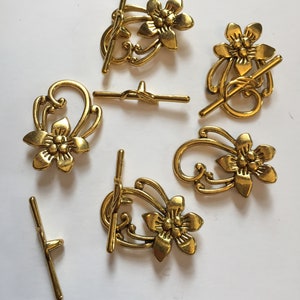 May include: Several gold-colored toggle clasps with a floral design. The clasps have a T-bar and a decorative loop with a flower. The clasps are made of metal and are arranged on a white surface.