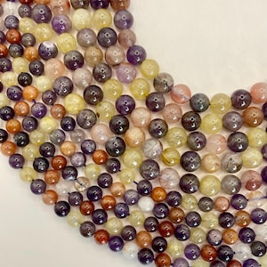 May include: Close-up of a collection of gemstone beads in various colors, including purple, yellow, orange, and clear. The beads are round and appear to be strung together, showcasing a variety of natural stone textures and hues, perfect for jewelry making.