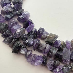 May include: Close-up of raw amethyst gemstone beads in shades of purple and gray. The irregular, rough-cut stones are strung together, creating a natural and textured appearance. These beads are suitable for jewelry making.