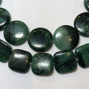 May include: Close-up of a necklace featuring large, polished green beads. The beads are a mix of round and square shapes, strung together with a visible string. The beads have a dark, mottled appearance with variations in color.