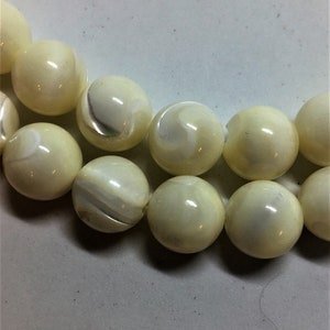 May include: Close-up of a strand of cream-colored, round beads. Each bead has a glossy finish with subtle marbling and variations in tone. The beads are strung together, suggesting a necklace or bracelet.