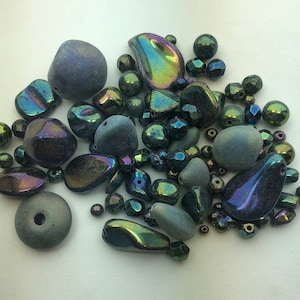 May include: A collection of various beads in shades of blue, green, and iridescent colors. The beads vary in shape and size, including round, faceted, and irregular forms. Some beads have a matte finish, while others are shiny.