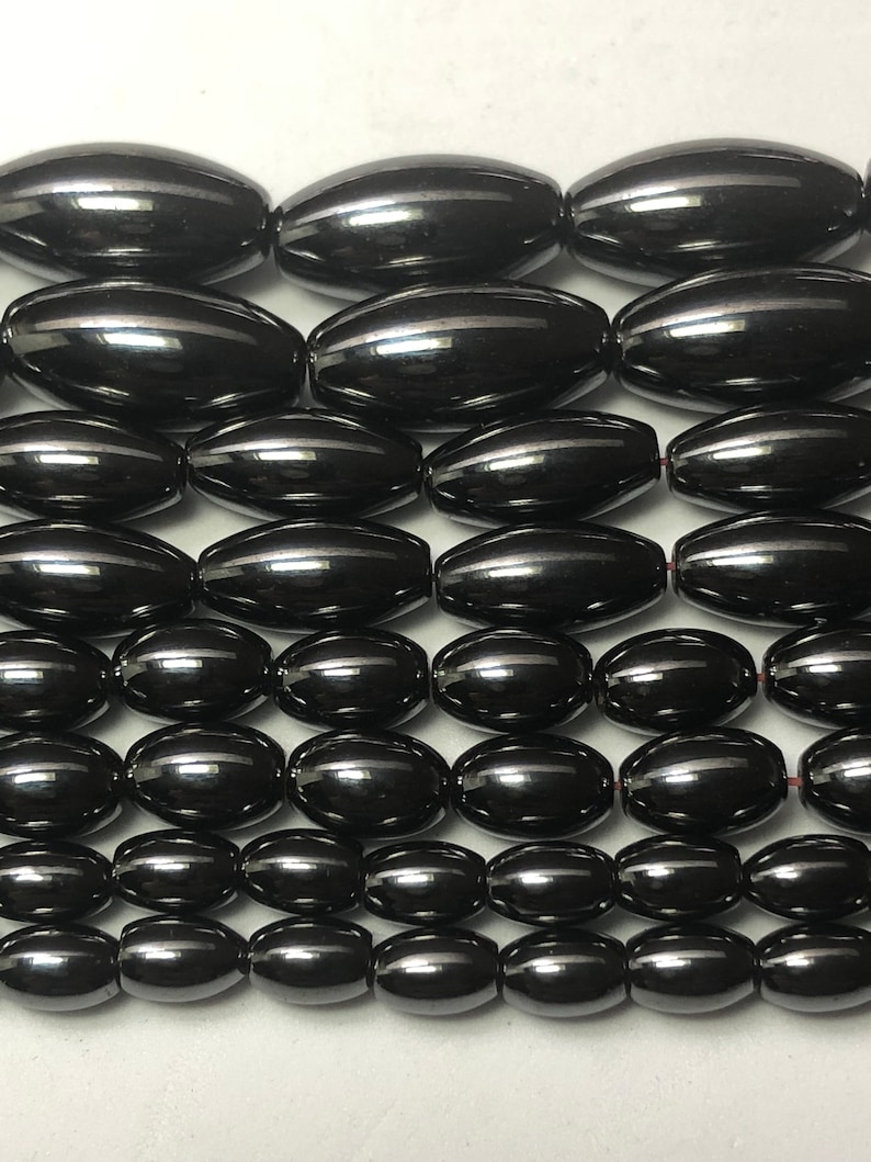 Peut inclure: Gros plan sur des rang&eacute;es de perles noires ovales et brillantes. Chaque perle pr&eacute;sente une surface r&eacute;fl&eacute;chissante avec une fine rayure claire. Les perles sont enfil&eacute;es, probablement pour la cr&eacute;ation de bijoux ou d'artisanat.