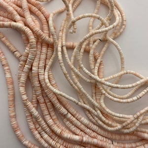 May include: Close-up of strands of small, flat, round beads in shades of cream and pale peach. The beads are strung together, creating a textured, organic pattern. The beads are likely used for jewelry making or other craft projects.