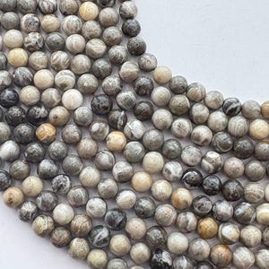 May include: A close-up view of a collection of round, natural stone beads. The beads vary in color, including shades of gray, cream, and brown, with unique patterns and textures. The beads are arranged closely together, suggesting they are strung or ready for jewelry making.