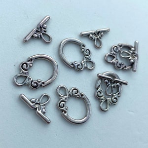 May include: A collection of silver-toned toggle clasps for jewelry making. The clasps feature a decorative design with a circular loop and a bar that fits through the loop. The clasps are in various sizes and orientations.